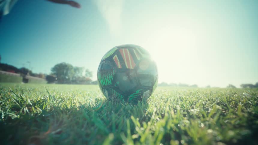 A runners legs striding powerfully kicks a soccer ball across dewbright green grass under a clear blue sky with sunlight flaring