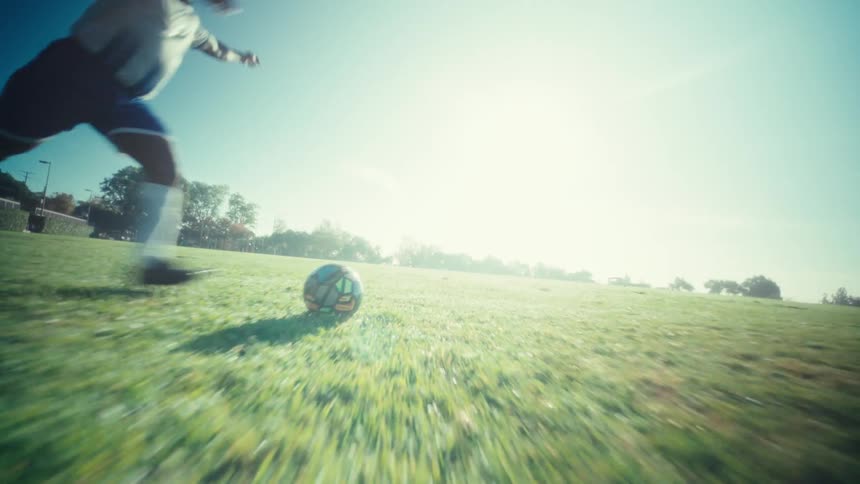 A man wearing a white soccer jersey with the number nine and blue shorts kicks a ball across a green grass field toward bright sunlight under a clear blue sky
