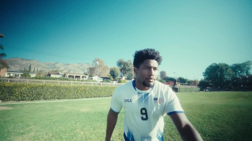 A man wearing a white soccer jersey with the number nine and blue shorts kicks a ball across a green grass field toward bright sunlight under a clear blue sky