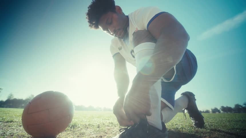 Man ties his shoe while half kneeling in a field near a soccer ball