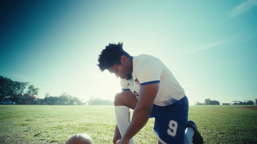 Soccer player kneeling to tie a cleat beside the ball on a grass field under sunlight