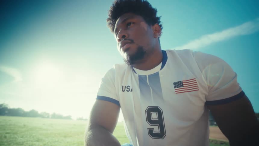 A young man in a usa soccer jersey sits on a sunny field looking down pensively before lifting his head to gaze into the distance with a serious determined expression against a bright clear sky