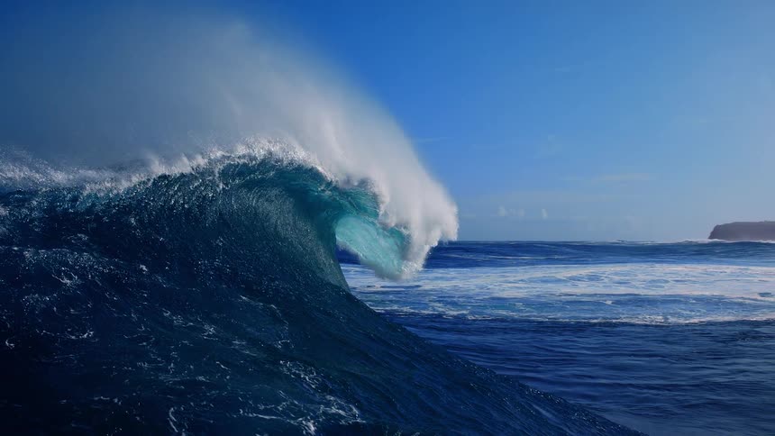 A wide view of a large ocean wave cresting and breaking into white foam under a clear blue sky with a rocky coastline visible in the distance