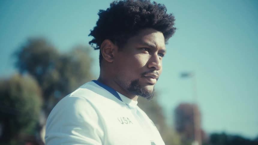A man wearing a white usa soccer jersey with the number nine looks up and sighs while standing on a grassy field under a clear blue sky on a bright day with a slight lens flare