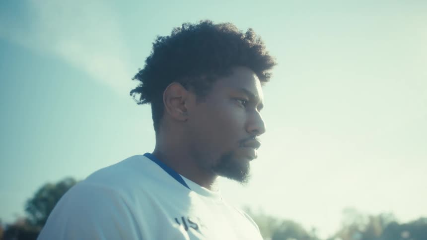 A man wearing a white usa jersey with the number nine stands outdoors under a clear blue sky the camera pans around him as he looks off into the distance eventually positioning the bright sun directly behind his head to create a heavy lens flare