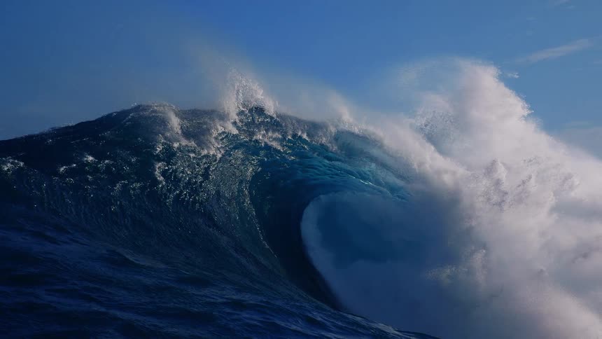 A massive ocean wave crests and breaks in the open sea sending thick white foam and spray into the air as the deep blue swell collapses under a clear sky