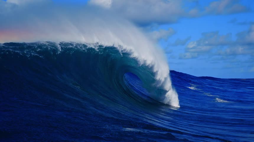 Powerful dark blue ocean waves swell and break into a hollow tube with white sea spray under a bright blue sky with scattered clouds