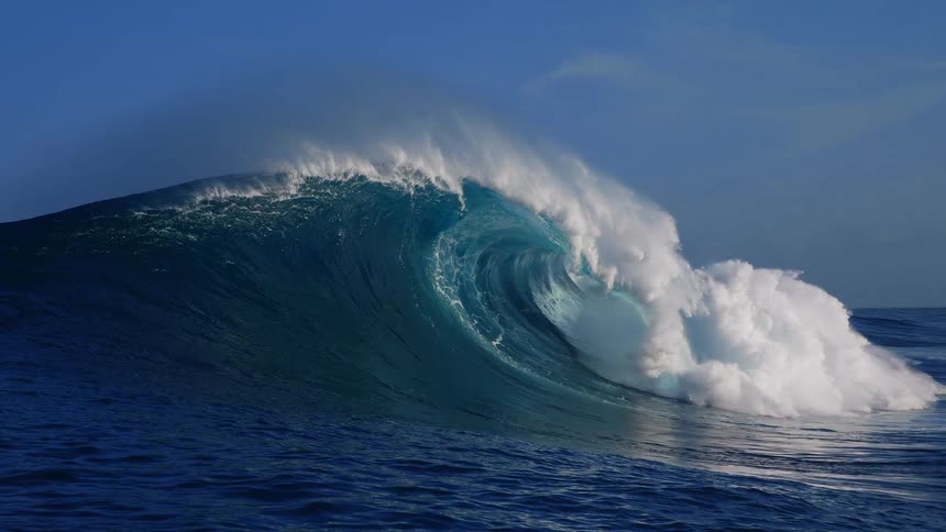 A massive ocean wave curls and crashes under clear blue sky