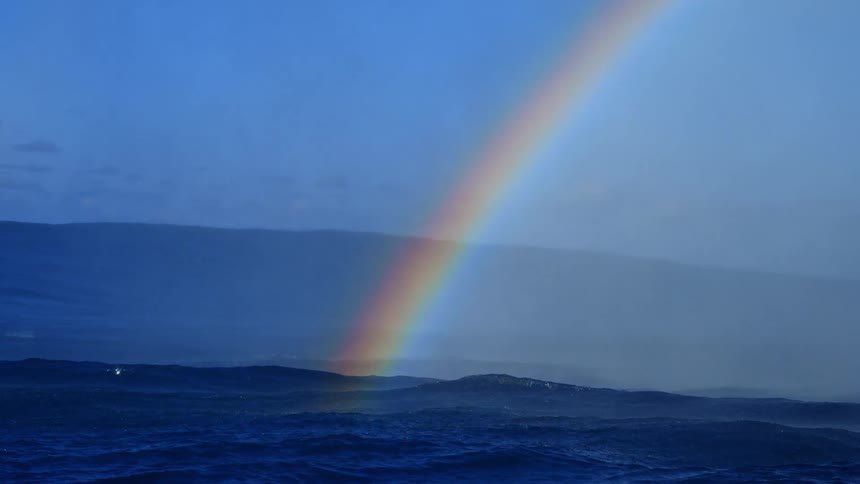 Waves splash as they roll in to shore in the rain with a rainbow stretching across the water