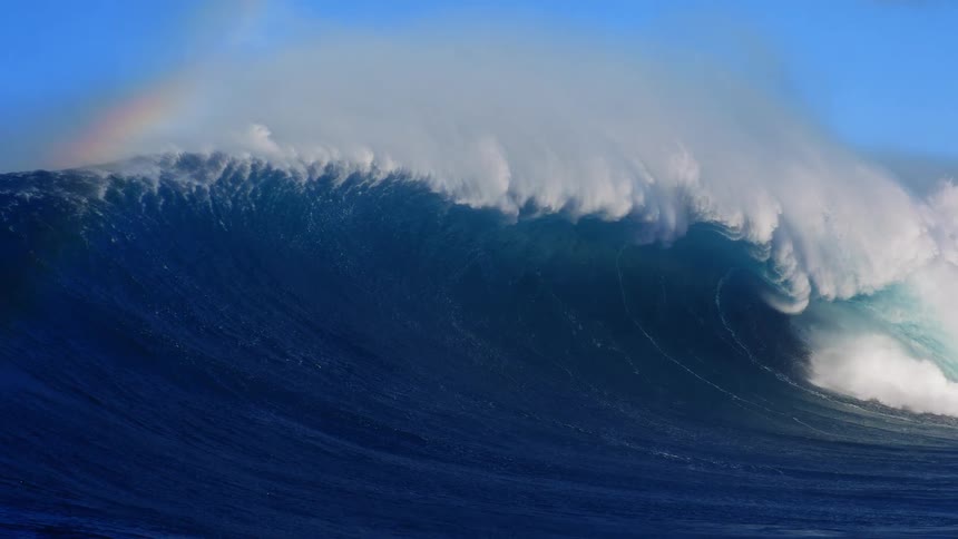 A faint rainbow appears over a massive dark blue ocean wave as it curls and breaks with white foam under a clear blue sky