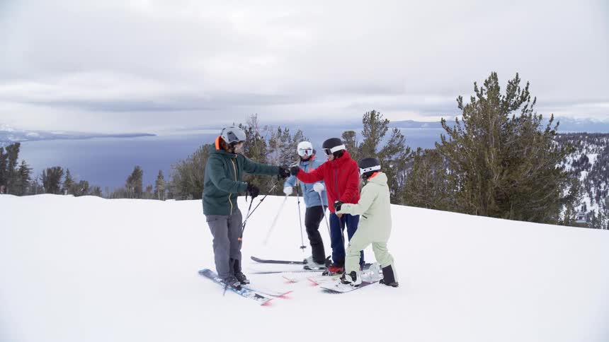 Two skiers standing on a snow covered mountain overlooking the coast as two other skiers join them