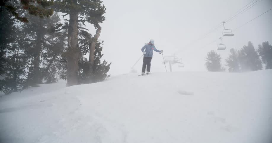 A person wearing a blue jacket and black pants skis down a snowy slope during a winter storm with a chairlift and evergreen trees visible in the foggy background