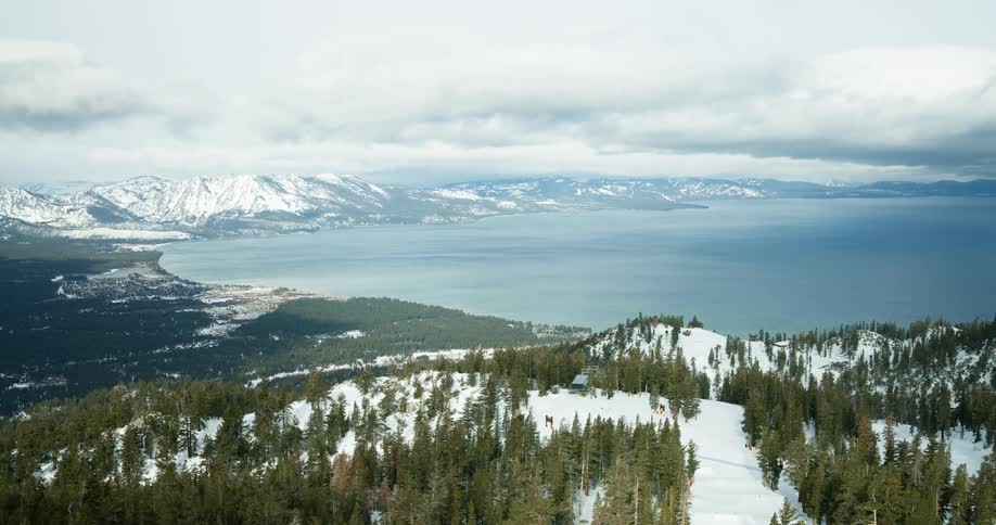 A mountain peak covered in snow and patches of short grass rises beneath a cloudy sky with the distant ocean visible in the background