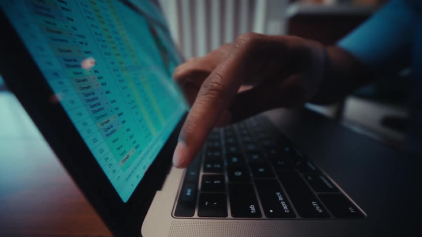 A finger taps a key on a laptop keyboard with a spreadsheet visible on the glowing screen
