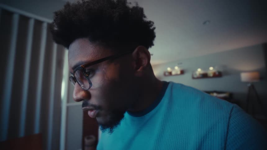 A man with glasses wears a blue shirt and sits at a wooden table in a dimly lit room intensely focusing on a laptop screen filled with spreadsheet data while his fingers move across the trackpad and keyboard to navigate the document