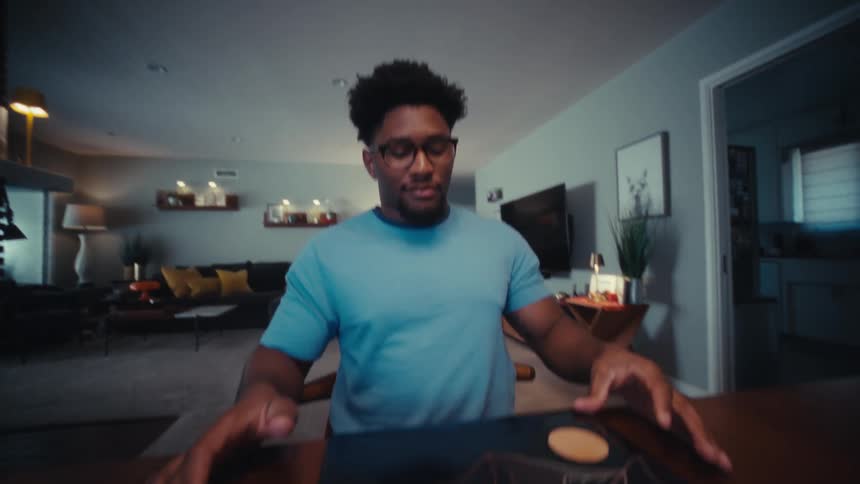 A man with glasses wearing a blue shirt closes his laptop while sitting at a wooden table in a living room at the end of a work session