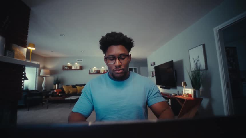 A young man wearing glasses opens his laptop at a table in a modern living room