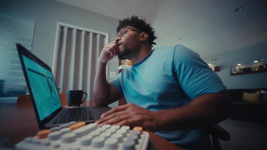 A young man sits in an office behind a desk with a computer wearing glasses as he works with steady focus