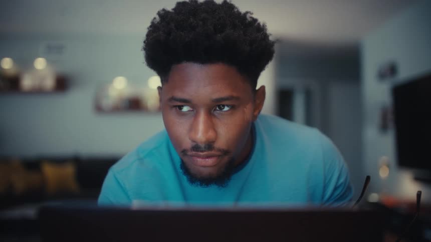 In a softly lit modern living room a focused black man wearing glasses and a blue tshirt sits upright at a table concentrating on his laptop