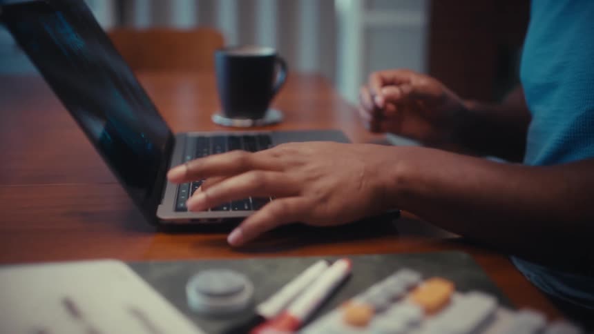 A person sets down a coffee mug opens a laptop and begins typing at a wooden table