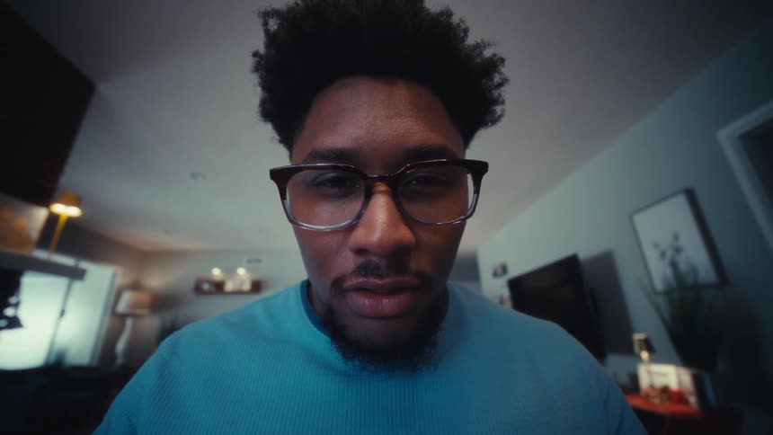 A young black male wearing a blue tshirt and clear glasses sits behind a computer typing with steady focus