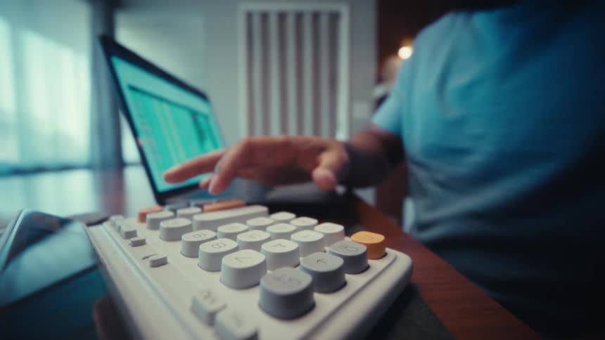 A man in a blue shirt uses a white calculator while working on a laptop with a financial spreadsheet in a modern home office