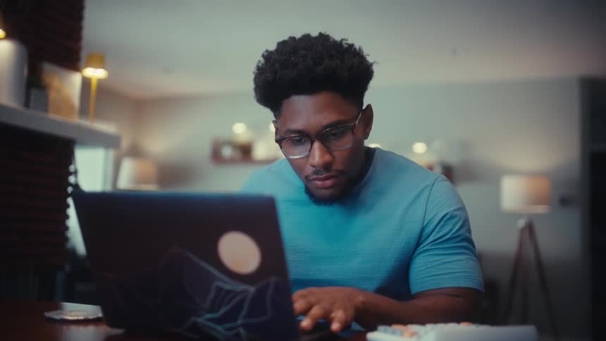 A man with glasses and a blue shirt works on a laptop and drinks from a black mug while sitting at a table in a cozy home office