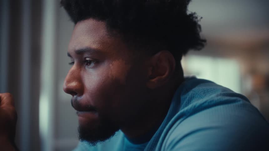 A young black male wearing a blue tshirt sits indoors in deep thought his expression calm and reflective as he focuses inward