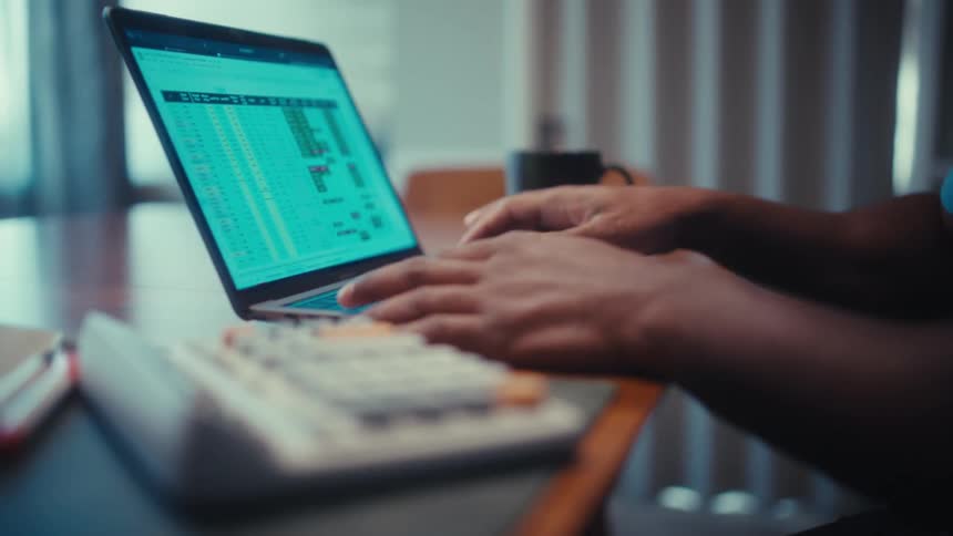 Black male person sitting and typing on a laptop computer with a calculating machine to the side over a desk indoors during day time