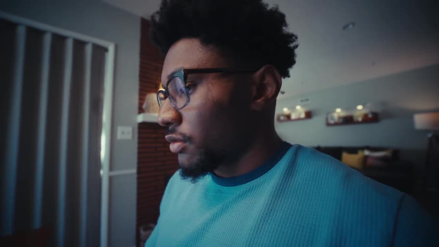 A young man wearing a blue tshirt and clear glasses sits behind a desk operating his laptop with focused attention