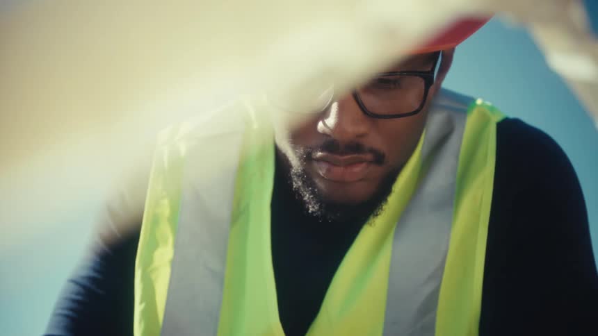 A focused engineer wearing a hard hat and safety vest works outdoors as the blades of a wind turbine model spin in the foreground