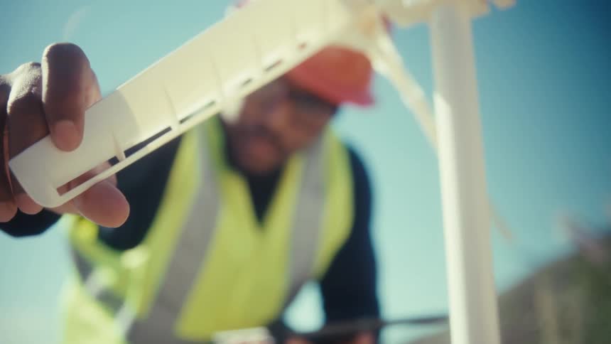 An engineer checks a tablet while inspecting a wind turbine at a sunlit windfarm