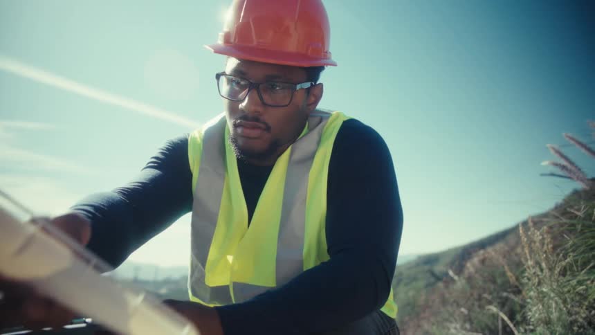 An engineer in a hard hat and safety vest uses a tablet to inspect a model of a wind turbine in the field