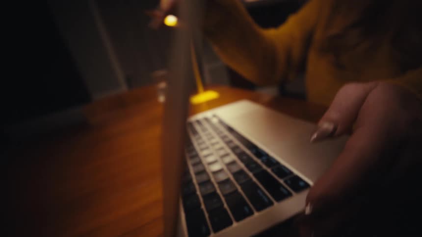 A young woman wearing a yellow sweatshirt closes the lid of her laptop on the desk a lamp glowing softly beside her