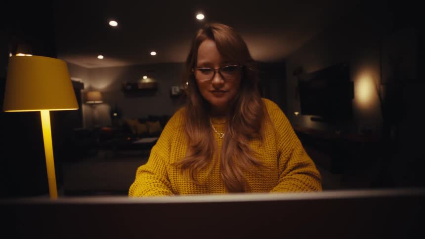 A young woman wearing a yellow sweatshirt sits behind a desk opening her laptop lid as a lamp glows softly beside her