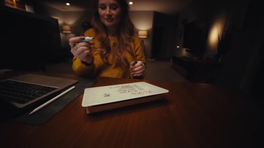 Close up of a young woman working with a laptop and marking a todo list while sitting at a table indoors at night