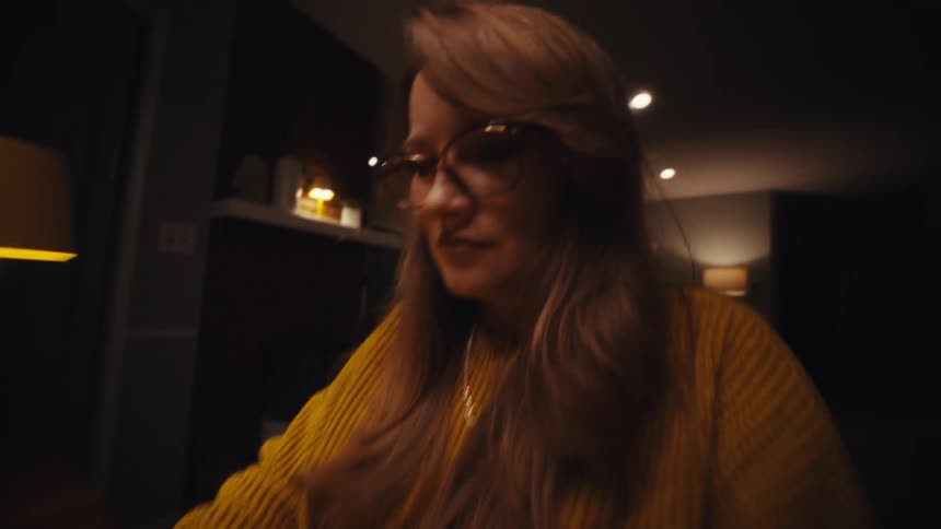 A young woman sits in a dark room behind a desk wearing glasses as she types on her laptop a glowing lamp beside her casting warm light across the space