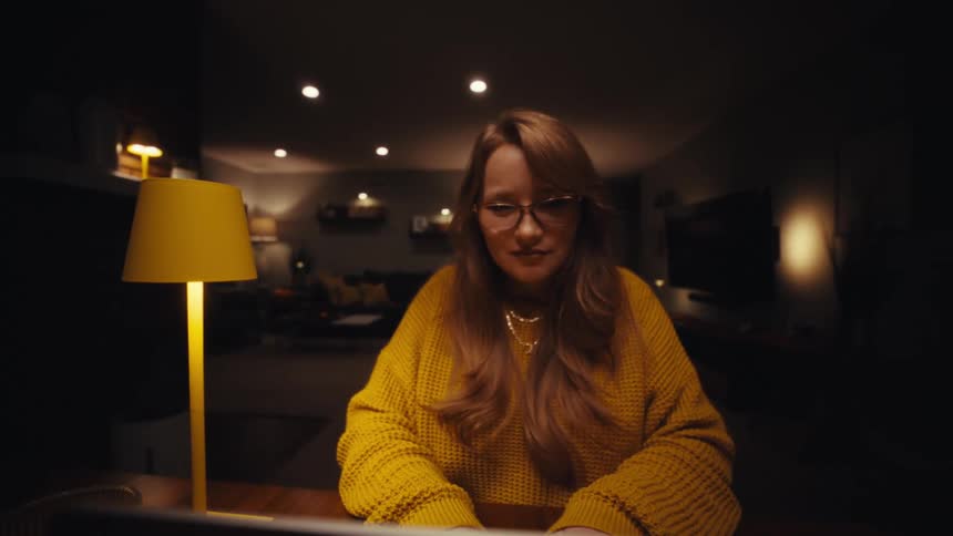 A woman types on her laptop at a wooden table illuminated by a yellow lamp in a dim room
