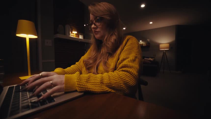 A woman wearing a yellow sweater and glasses opens her laptop and begins typing while sitting at a wooden table in a dimly lit room with a warm lamp
