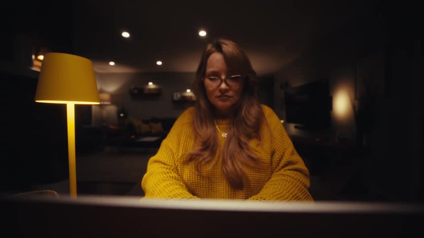A woman in a yellow sweater opens her laptop at a desk in a dimly lit room to begin working