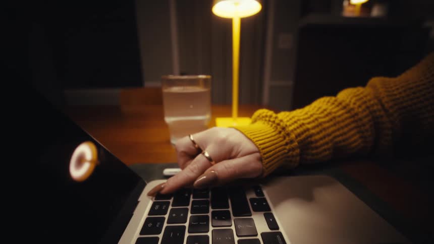 Close up of a woman putting on her laptop computer while sitting at a table indoors at night