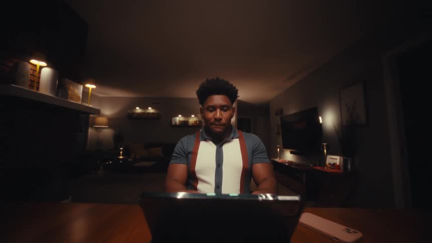 A man wearing a polo shirt with suspenders sits at a table in a dimly lit room and looks at a tablet screen before leaning forward