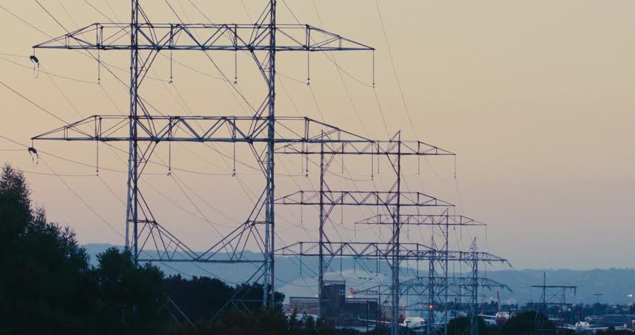 A line of tall steel transmission towers carries electrical wires across a valley toward a distant city during a hazy sunset while cars drive on a highway below