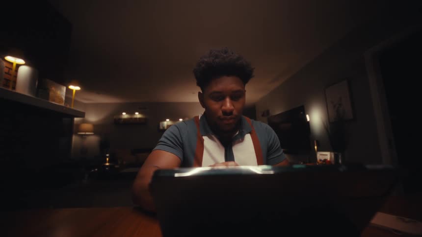 A man with short curly hair and a patterned polo shirt types on a white keyboard and uses a tablet while working at a desk in a dark room at night