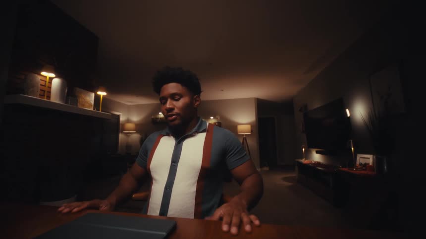 Young adult black man sitting down in front of a desk closing a tablet computer and reclining on the chair with a relieved facial expression indoors during night time
