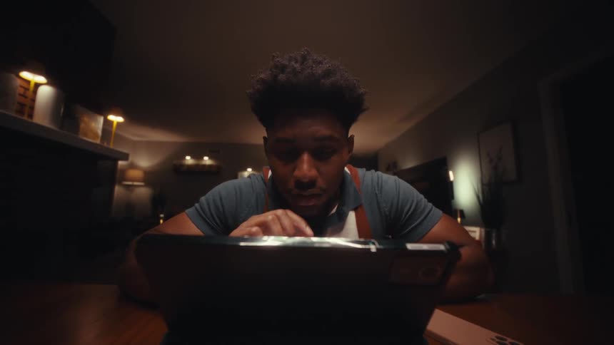 A focused young man in suspenders sits at a wooden table in a dimly lit room intensely interacting with a laptop while a smartphone lies nearby in the background