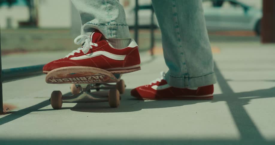 A person wearing red sneakers and light blue jeans places their front foot on a skateboard and pushes off the concrete pavement to begin riding
