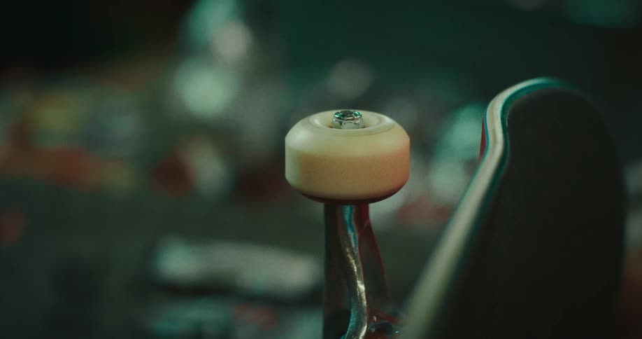 A persons hand reaches into the frame and spins a white wheel attached to a skateboard truck which continues to rotate rapidly against a blurred workshop background