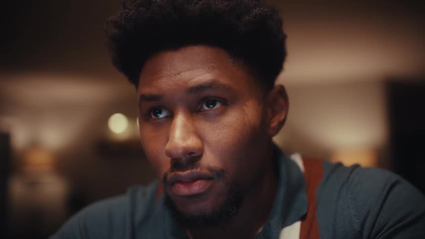 Close view of a man wearing a blue shirt and an apron looking down with a focused expression in a room with warm soft lighting in the background