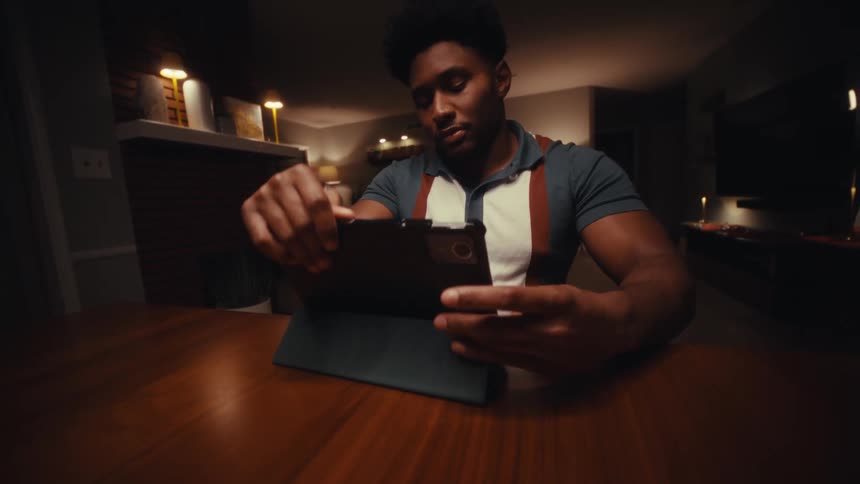 A man wearing a patterned polo shirt opens a tablet computer with a grey cover and starts typing while sitting at a wooden table in a dimly lit room at night